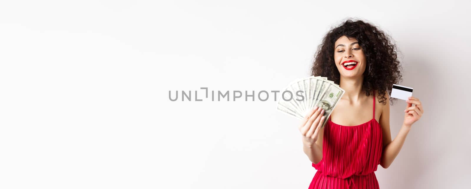 Shopping. Rich successful woman with curly hair and red dress, holding plastic credit card and looking pleased at dollar bills, white background.
