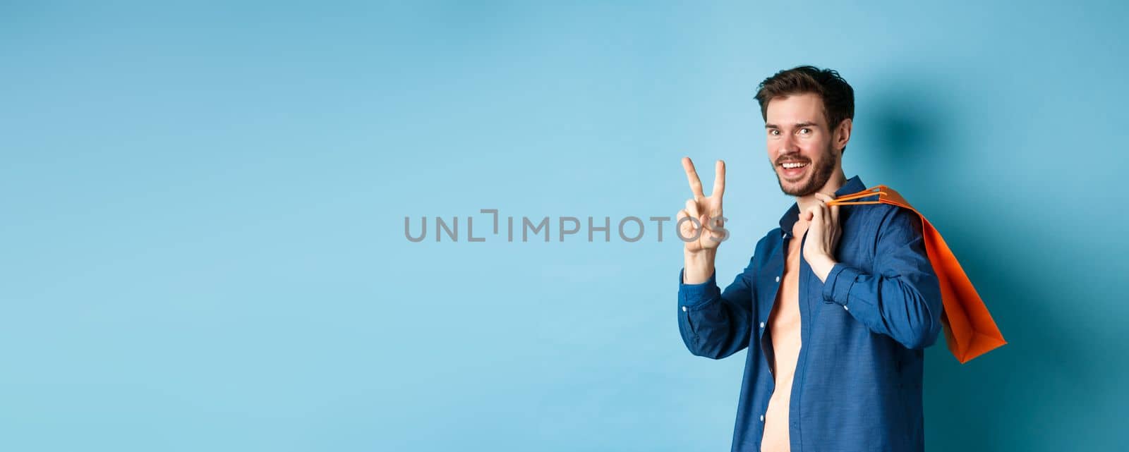 Cheerful guy holding orange shopping bag on shoulder, smiling and showing peace sign, standing on blue background. Copy space