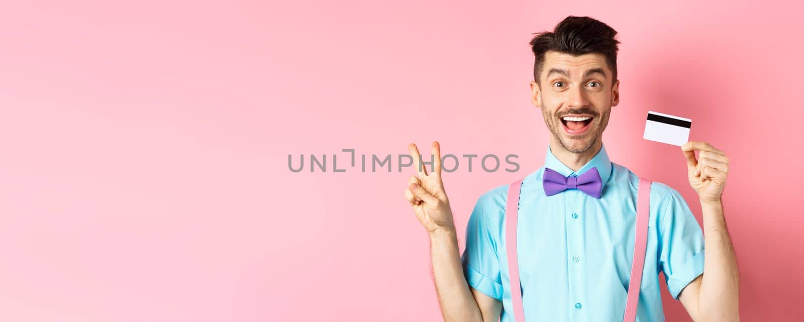 Shopping concept. Smiling caucasian guy in bow-tie showing plastic credit card and peace, victory sign, standing happy on pink background.