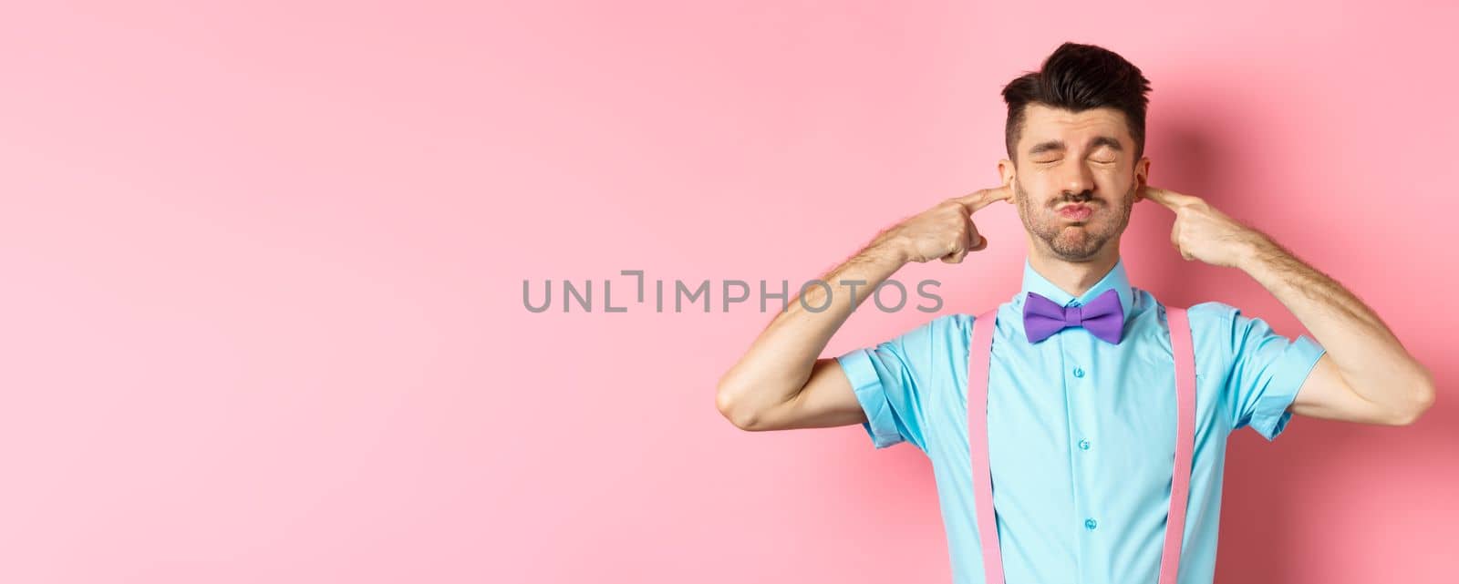 Annoyed funny guy in bow-tie shut ears, plug fingers and grimacing, irritated by loud noise, standing over pink background.