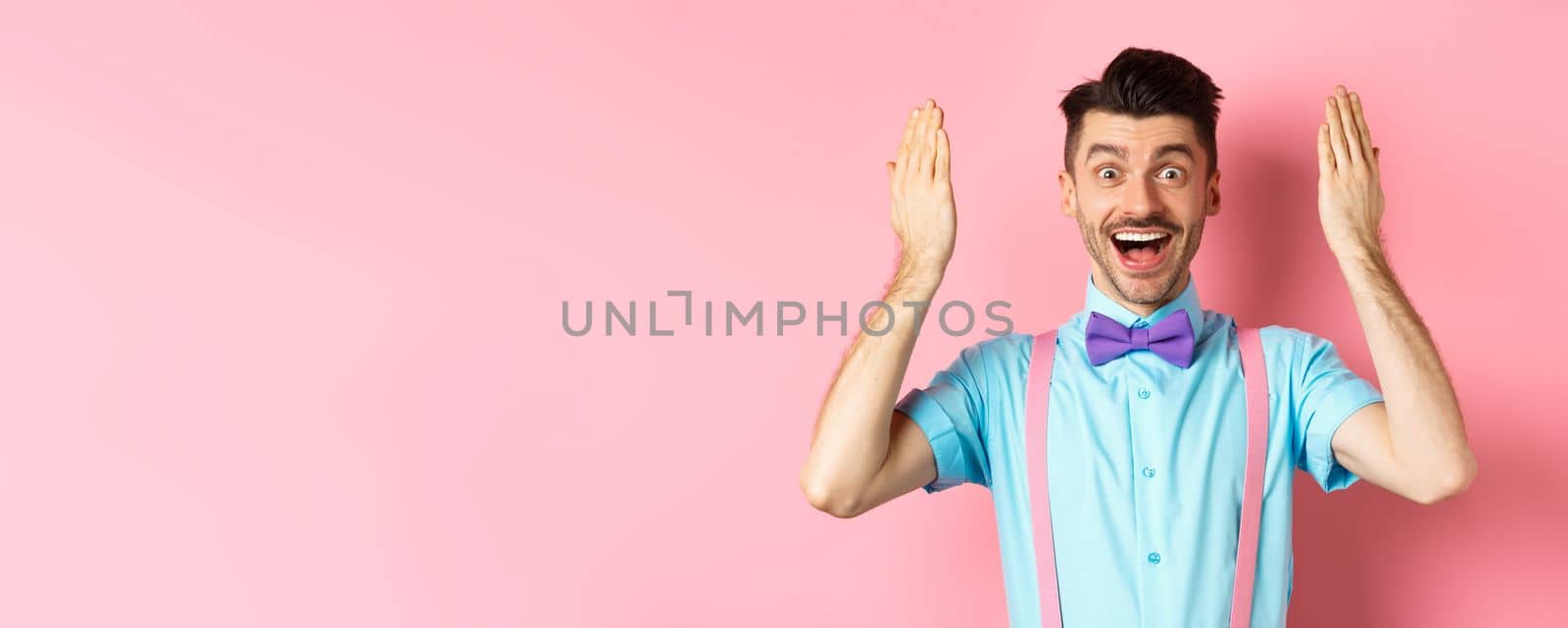 Cheerful young man open eyes for surprise present on holiday celebration, looking amazed at gift, standing on pink background.
