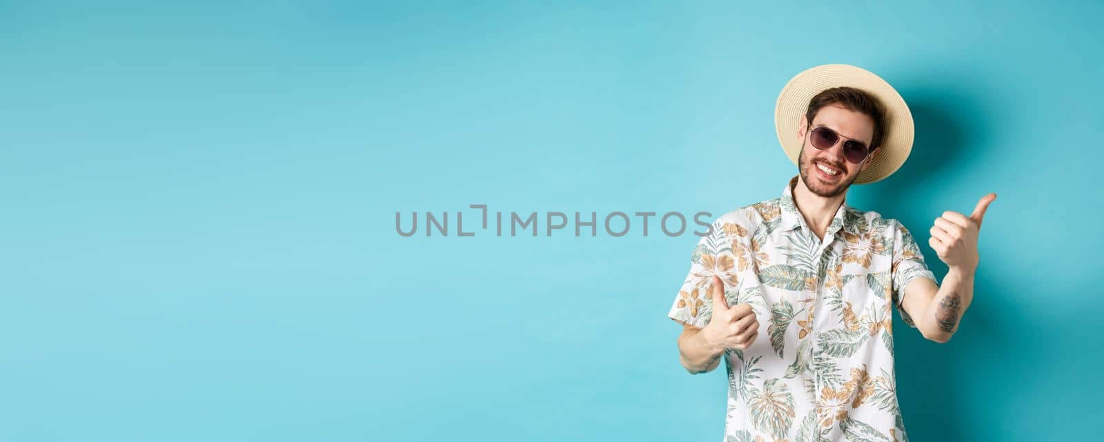 Cheerful tourist having fun summer holidays, showing thumbs up and smiling, standing in hawaiian shirt and sunglasses on blue background.