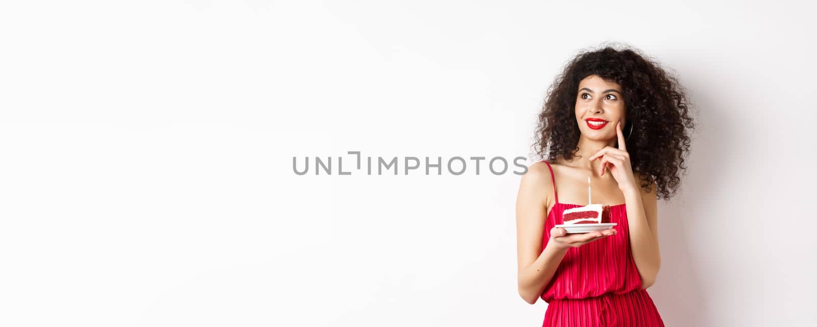 Happy woman celebrating birthday, holding cake and making wish, looking aside dreamy, standing in red dress on white background. Celebration and holiday concept.