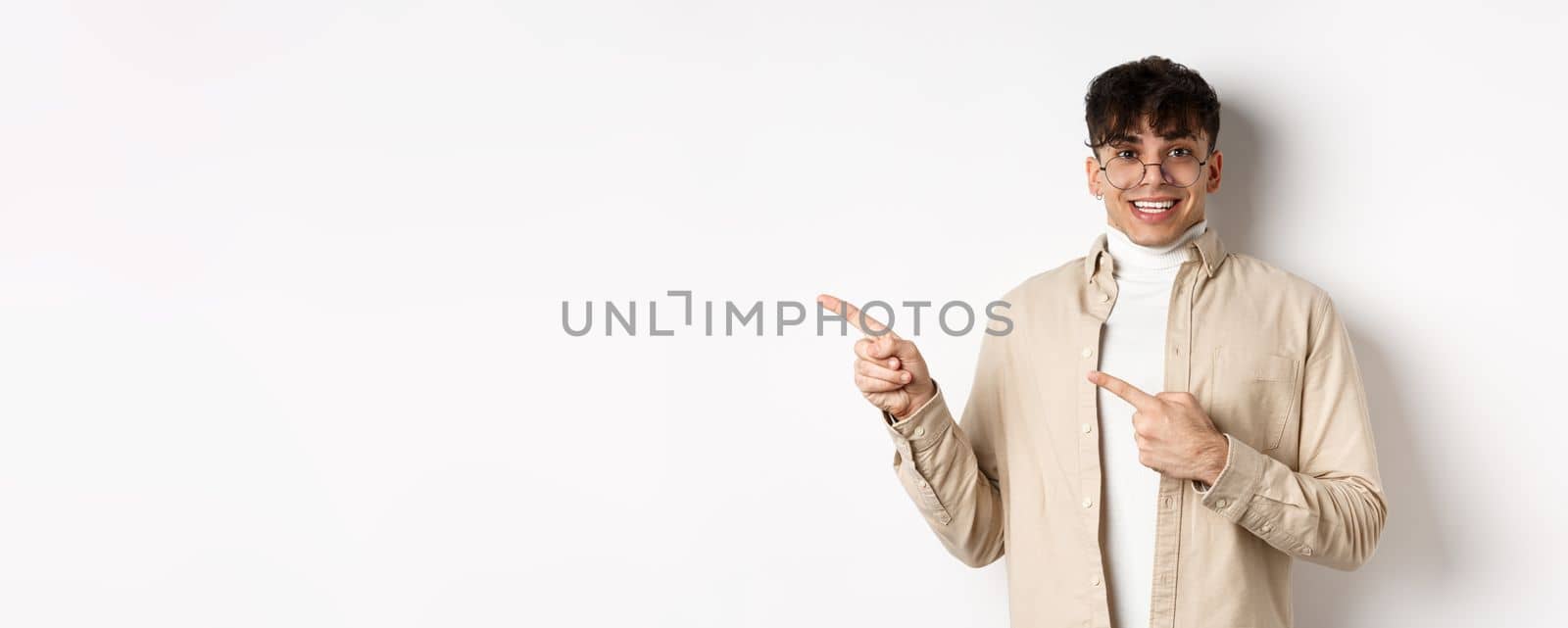 Cheerful handsome man in glasses smiling, pointing fingers left at logo, showing advertisement with excited face, standing on white background.