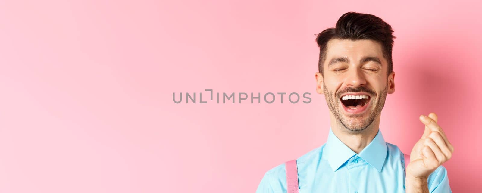Valentines day concept. Happy man in love showing finger heart and laughing, standing on romantic pink background by Benzoix