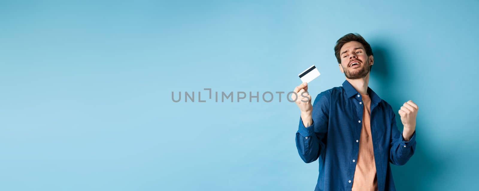 Happy and satisfied young man dancing with plastic credit card, fist pump and smile, standing on blue background.