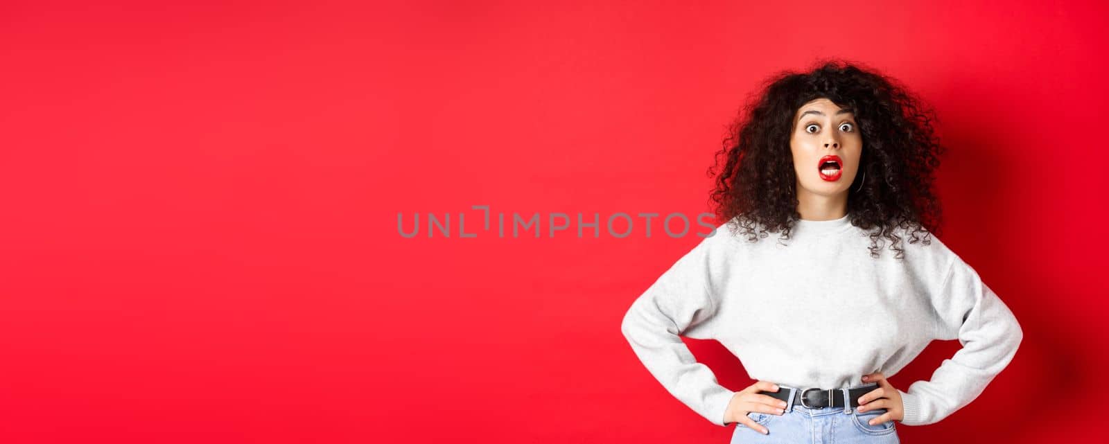 Shocked italian woman with curly hair, gasping and staring at camera astonished, open mouth, standing in white sweatshirt on red background.