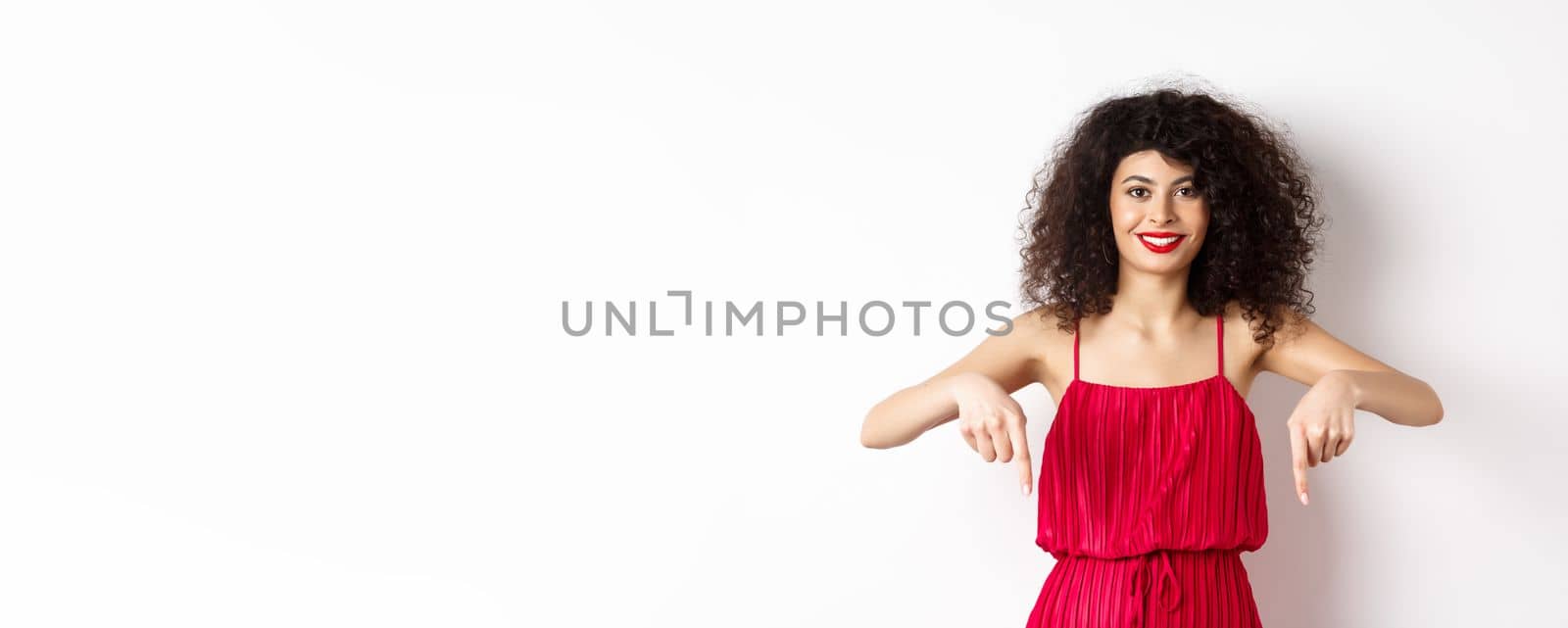 Attractive female model in red dress and makeup, pointing fingers down and smiling, showing advertisement, standing over white background by Benzoix