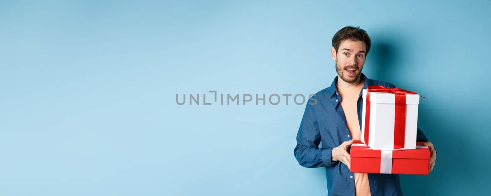 Handsome bearded guy holding Valentines day gifts for lover, standing with presents in boxes and looking at camera, blue background by Benzoix