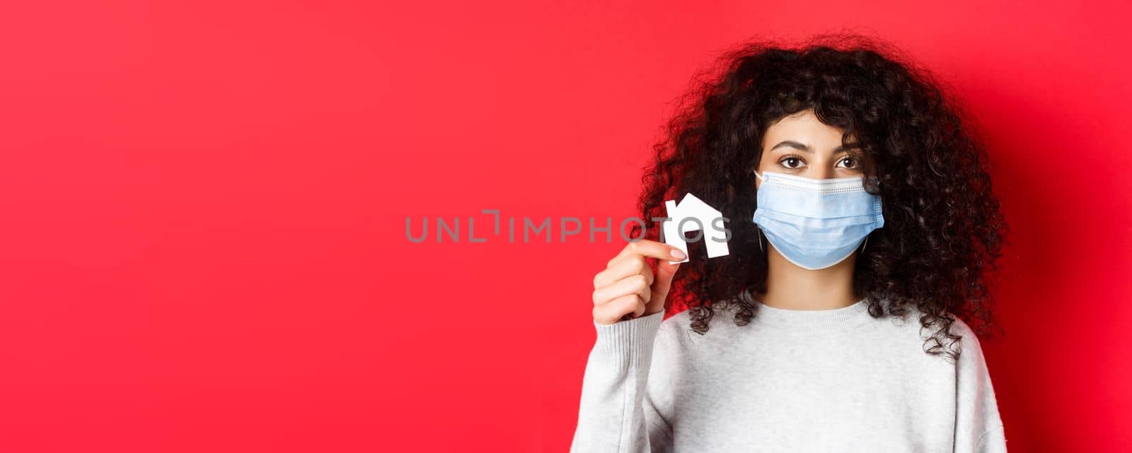 Real estate and pandemic concept. Young woman in medical mask showing small paper house cutout, standing on red background.