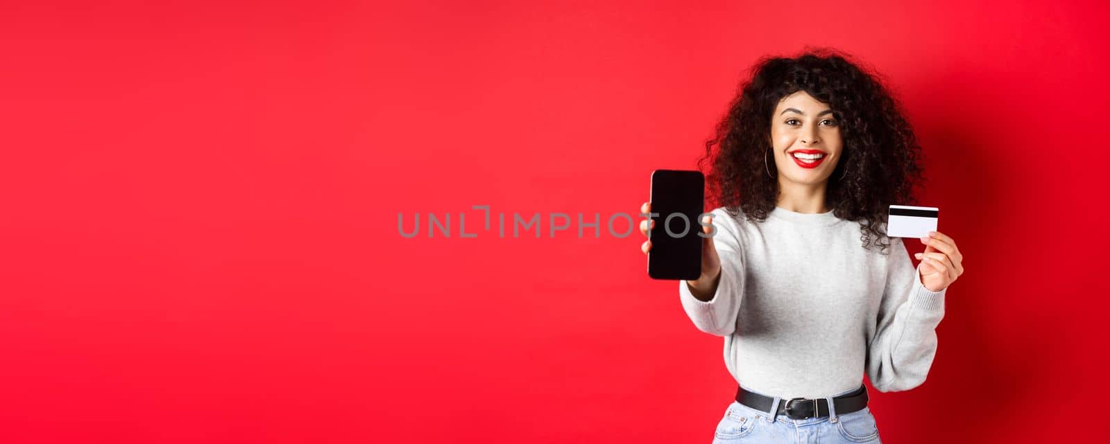 Happy good-looking girl showing plastic credit card and empty smartphone screen, demonstrate account or application, standing on red background.