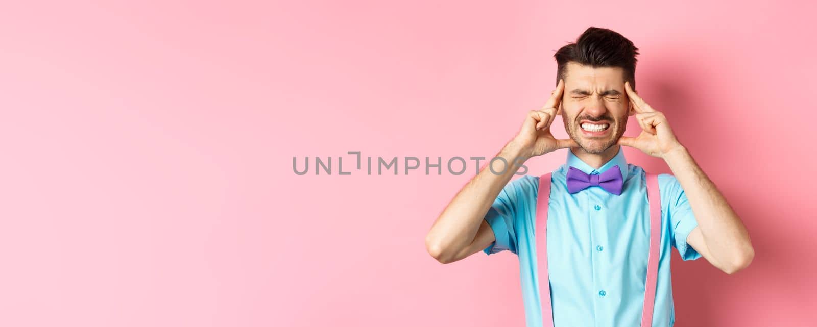 Image of man with headache touching head temples, grimacing from painful migraine, clench teeth, standing sick on pink background.