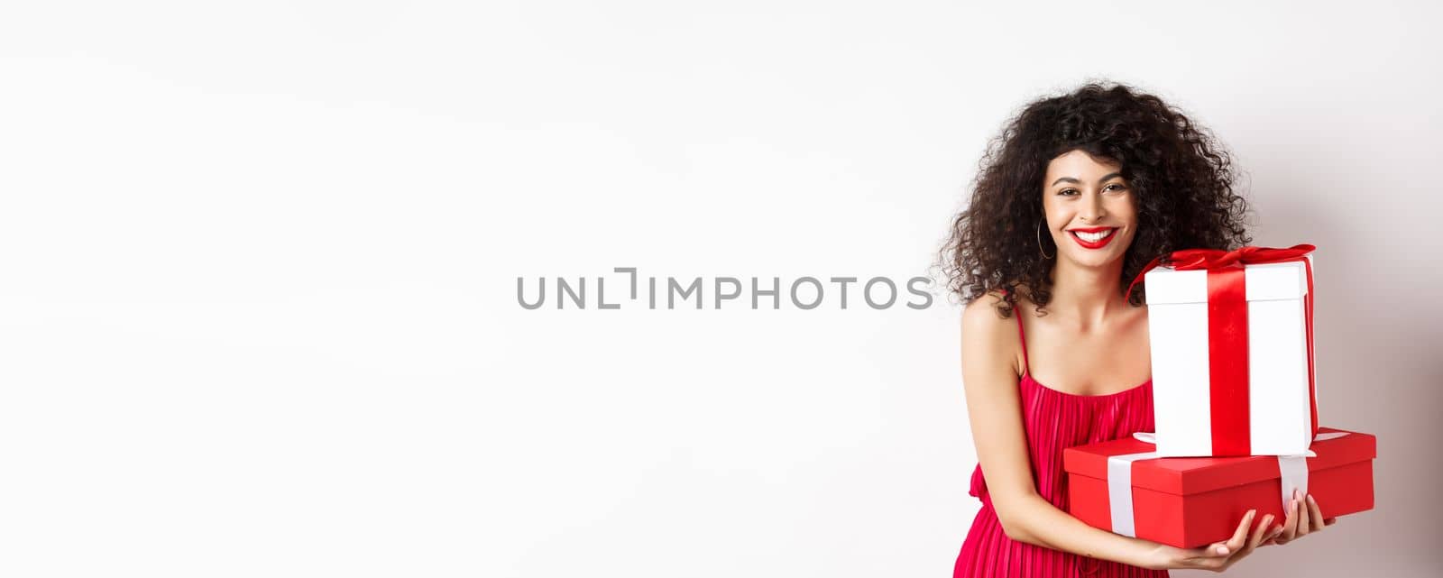 Beautiful birthday girl with curly hair, holding bday gifts and smiling happy, celebrating, standing against white background by Benzoix