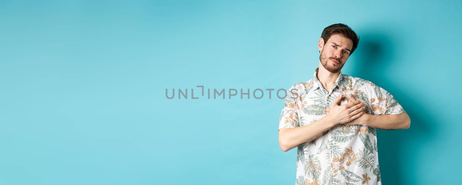 Passionate and romantic guy in hawaiian shirt, holding hands on heart and looking with sympathy, being in love, standing on blue background.
