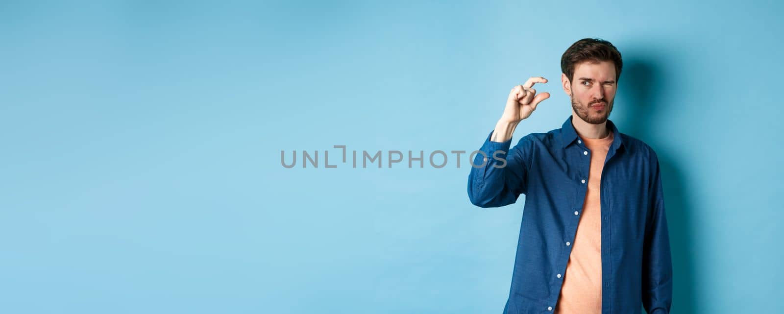 Image of young caucasian man showing small size and squinting to see something tiny, standing on blue background.