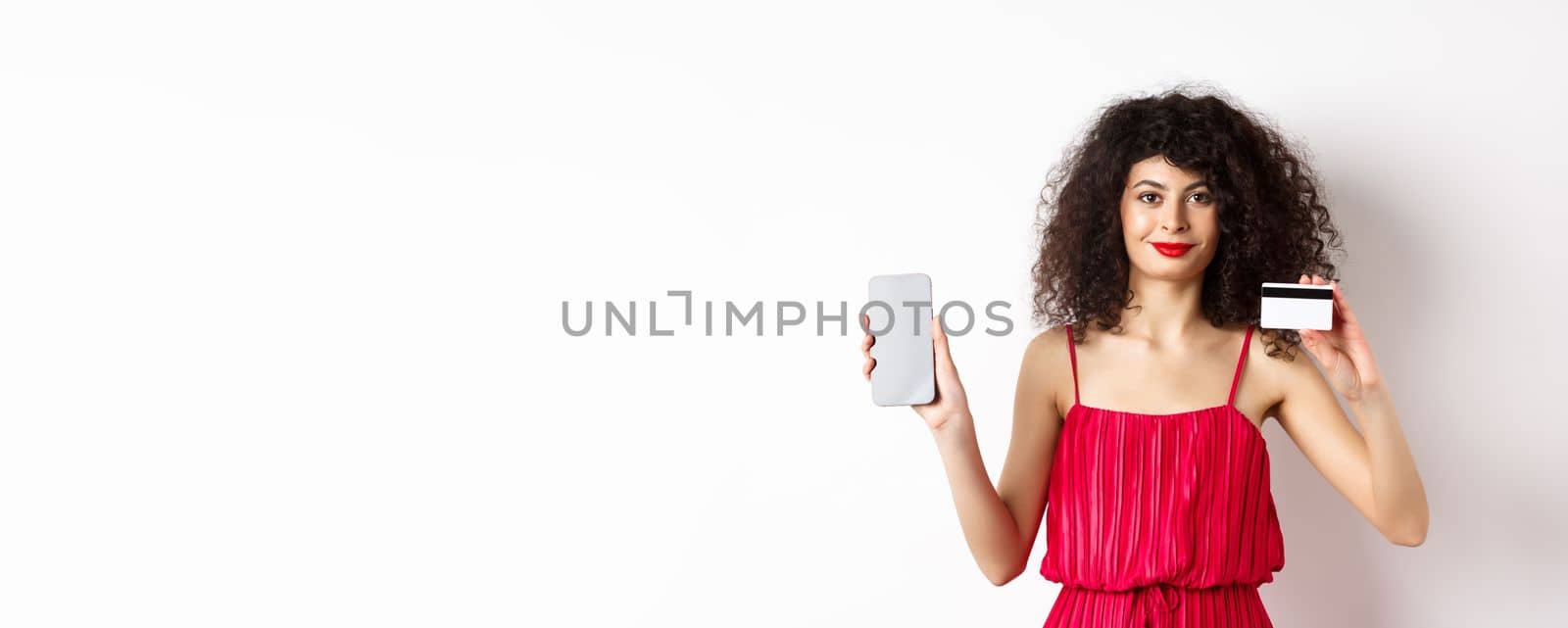 Online shopping. Fashionable woman with curly hair, wearing red dress, showing smartphone screen and plastic credit card, smiling at camera, white background.