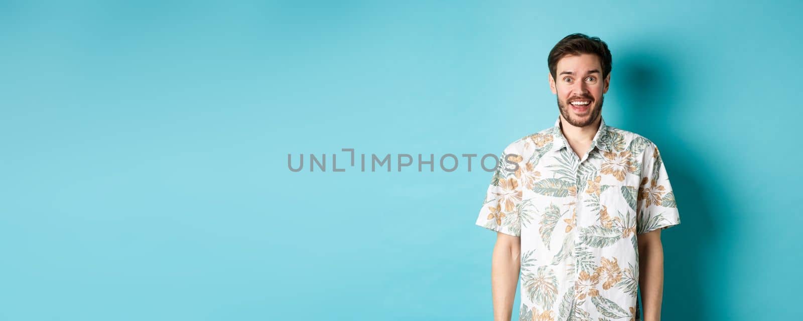 Summer holiday. Handsome happy man in hawaiian shirt looking amused, smiling at camera, standing on blue background.