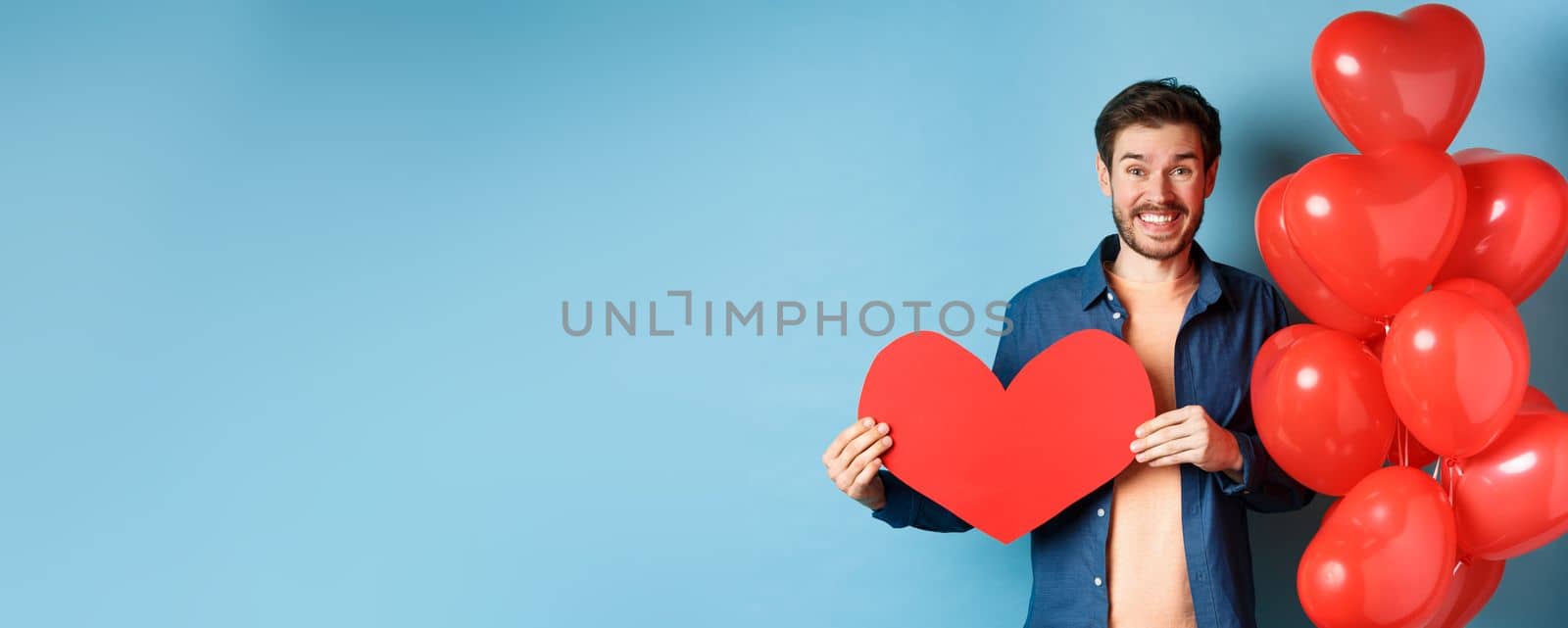 Valentines day concept. Smiling man say I love you, holding paper red heart cutout, standing near romantic balloons, blue background.
