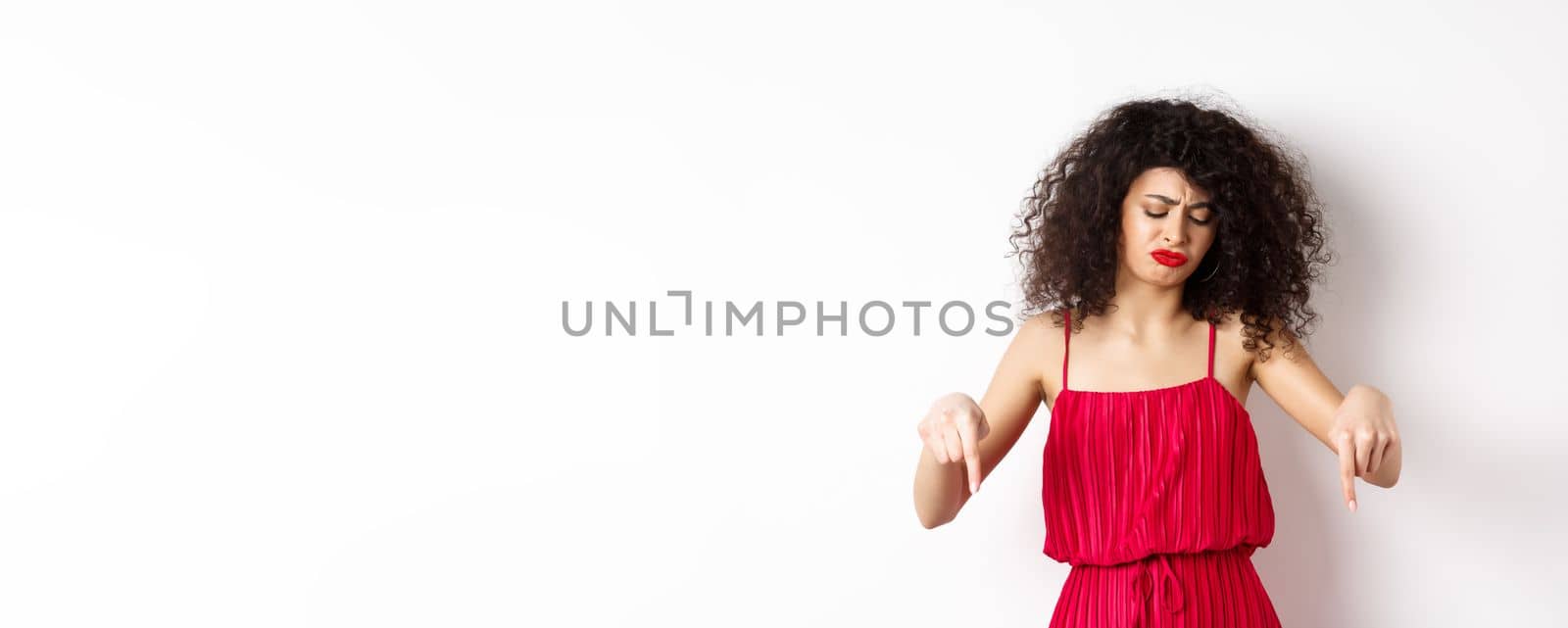 Sad and disappointed curly-haired woman in red dress, frowning and looking down jealous, pointing at promo, standing over white background by Benzoix