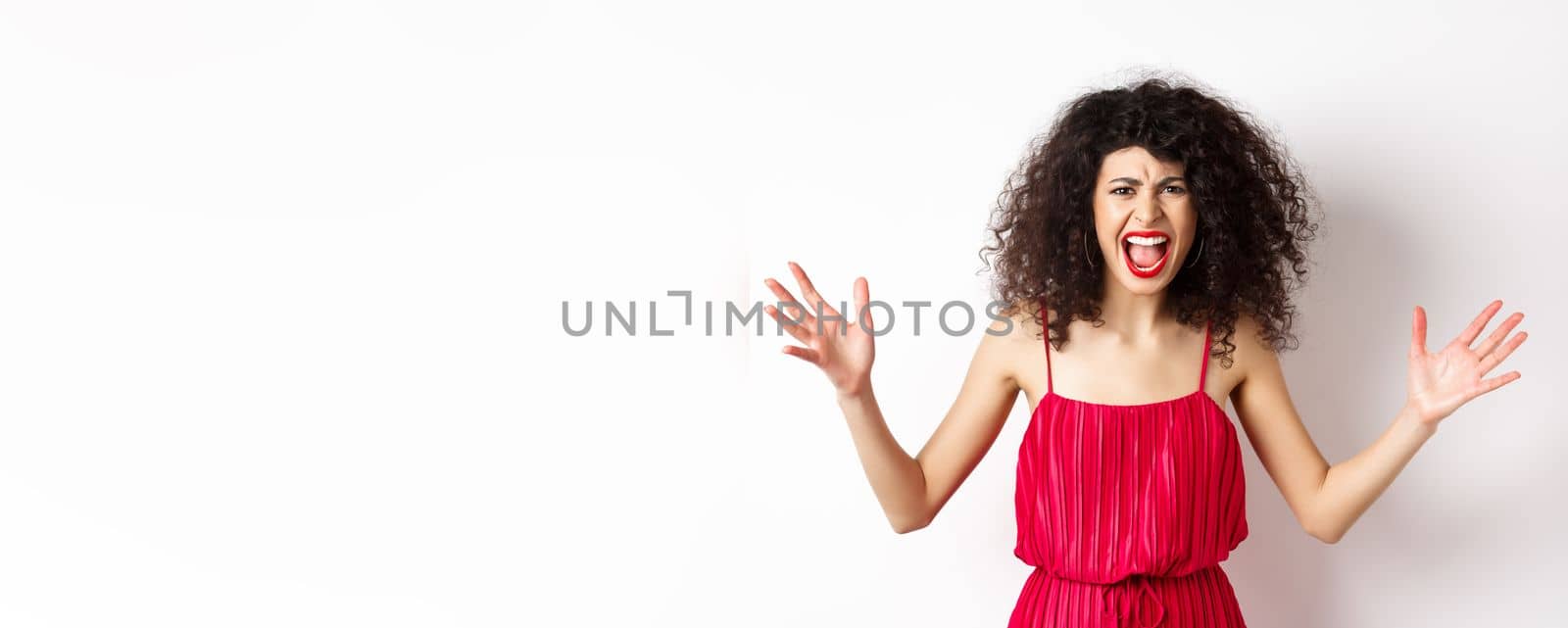 Angry young woman with curly hair, wearing red dress, screaming and having an argument, looking with hatred and anger, standing over white background.
