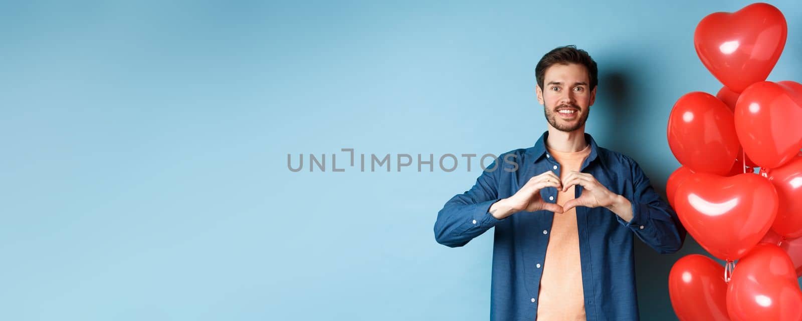 Happy valentines day. Boyfriend in love showing heart gesture to lover and smiling, standing near red balloons on blue background by Benzoix