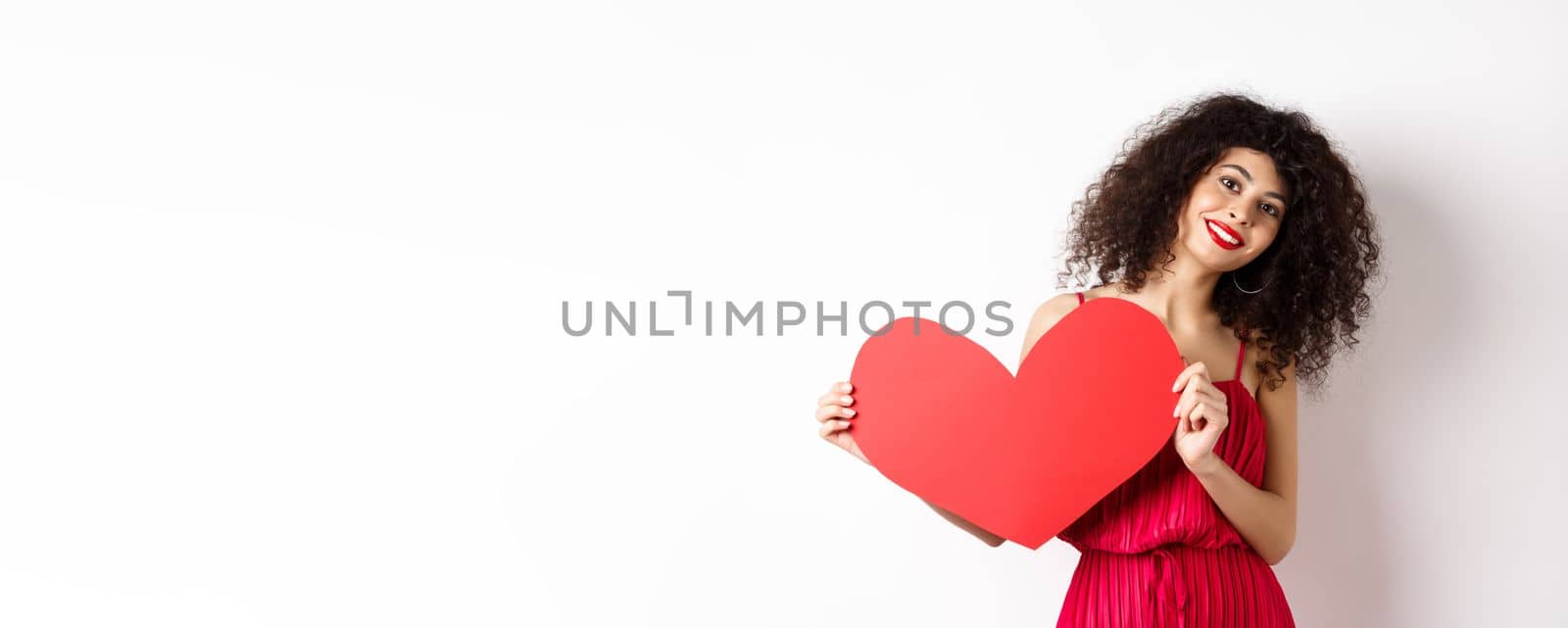 Romantic woman in dress showing big red heart, falling in love, smiling happy at camera, white background by Benzoix