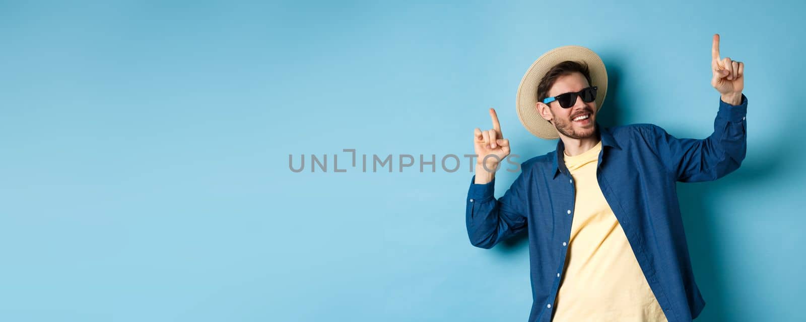 Funny tourist dancing on vacation, pointing fingers up, wearing summer hat and sunglasses, blue background.