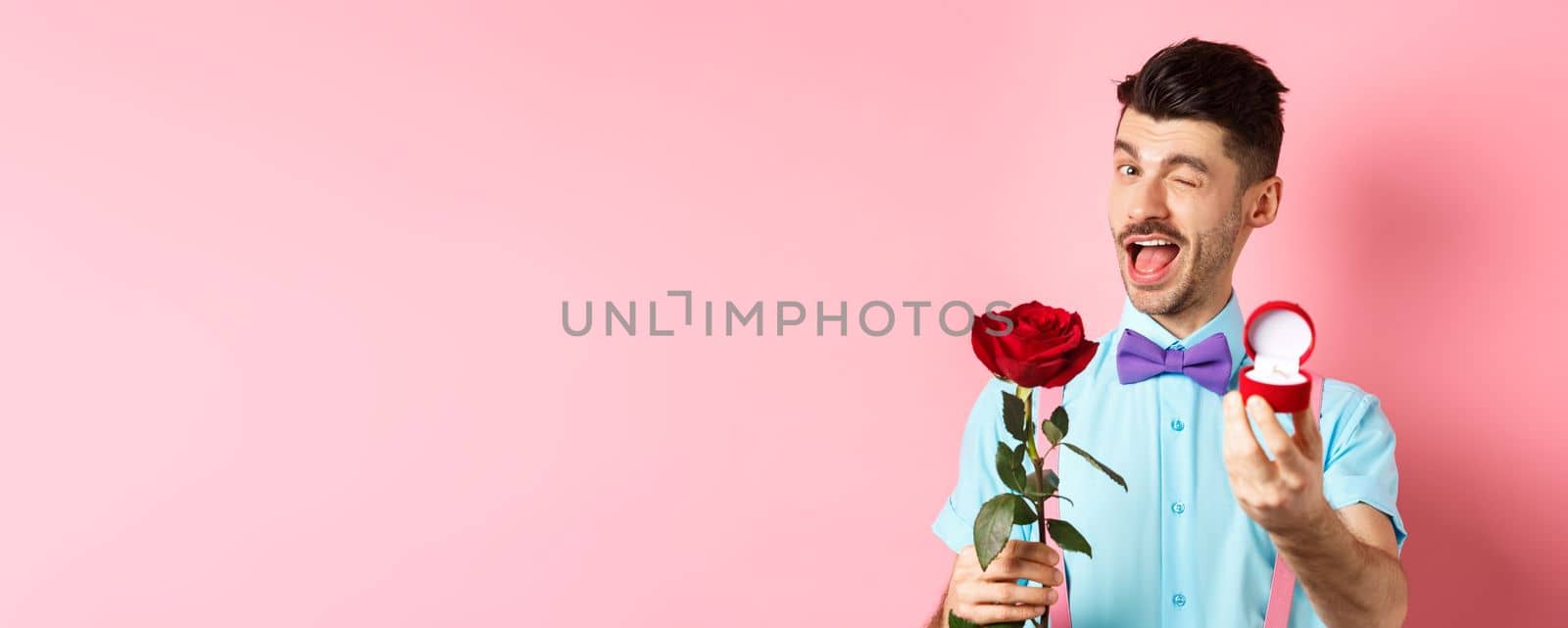 Valentines day. Funny guy making proposal, winking and saying marry me, showing engagement ring with red rose, standing over pink background.