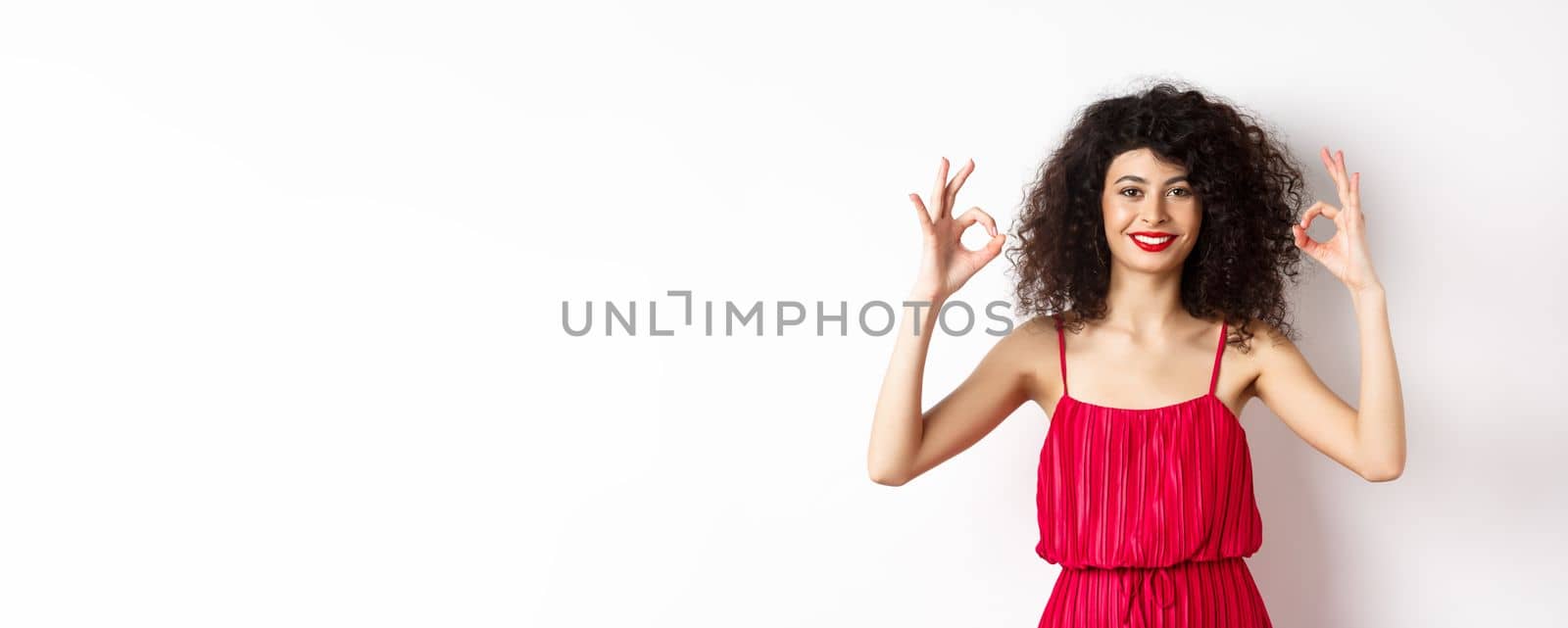 Beautiful woman in red dress, smiling and showing okay gestures, like something good, praising good job, standing over white background.
