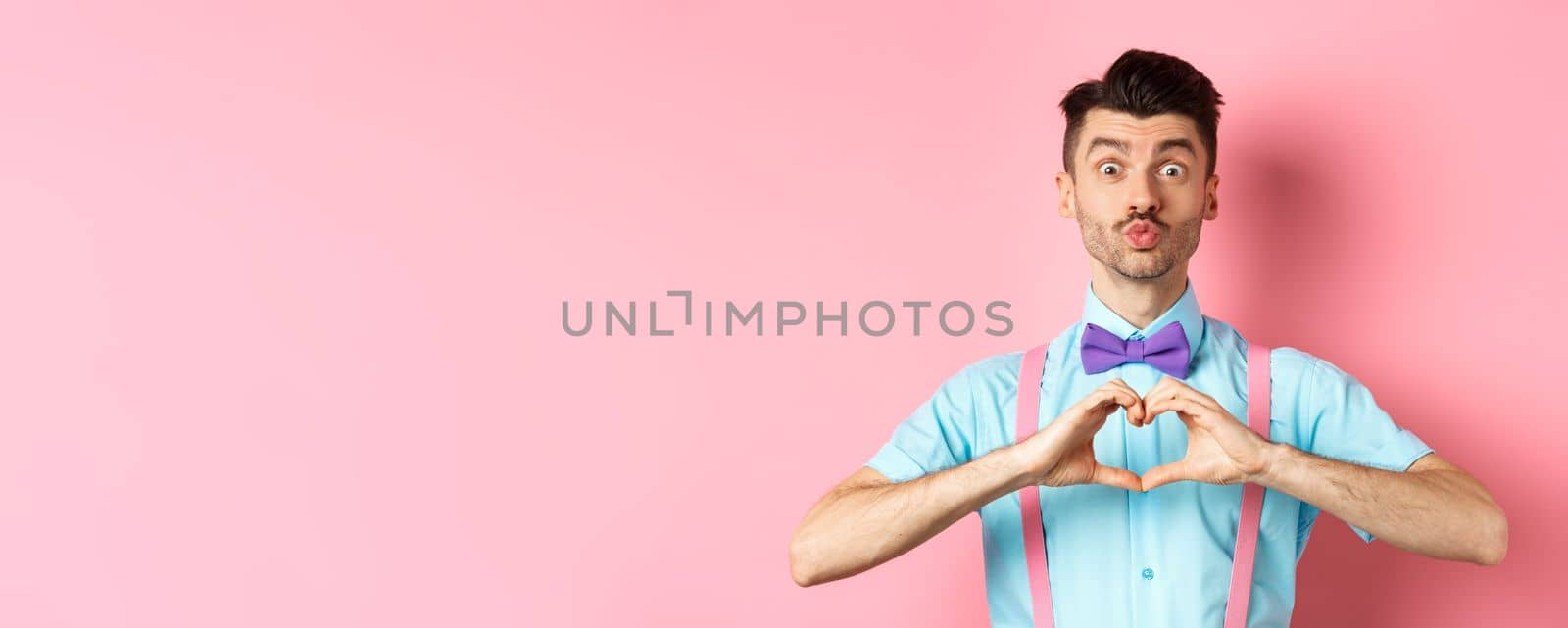 Happy Valentines day. Funny young man waiting for lovers kiss, pucker lips and show heart sign, I love you gesture, express feelings on date, pink background by Benzoix