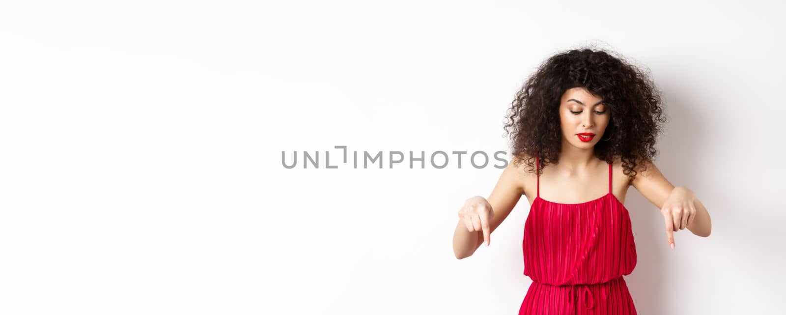 Intrigued beautiful woman with red lips and dress, pointing and looking down at banner, reading logo, standing over white background by Benzoix