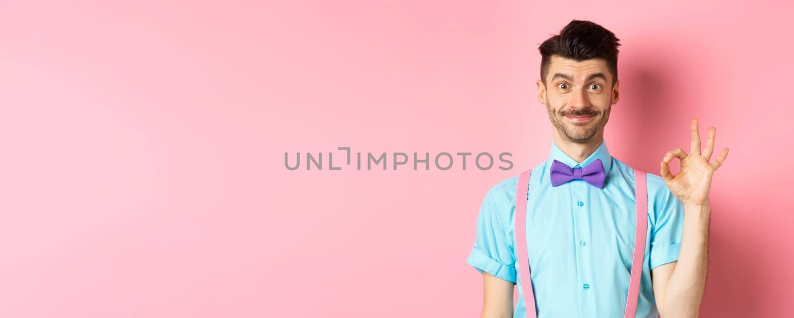 Smiling caucasian man in bow-tie and suspenders showing all okay gesture, recommending something cool, praising promo on pink background.