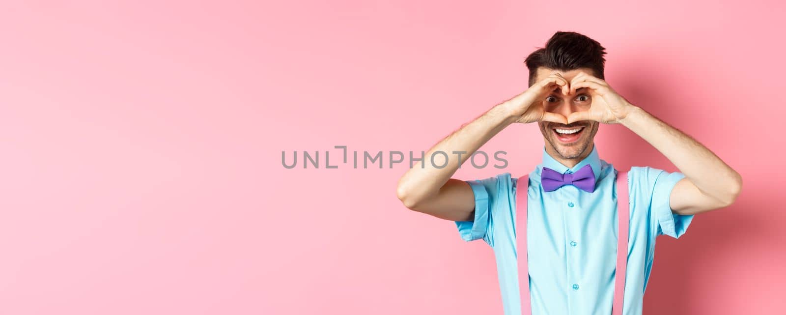 Happy Valentines day. Young man making hand heart and looking through it at camera, feeling romantic with lover, standing over pink background by Benzoix