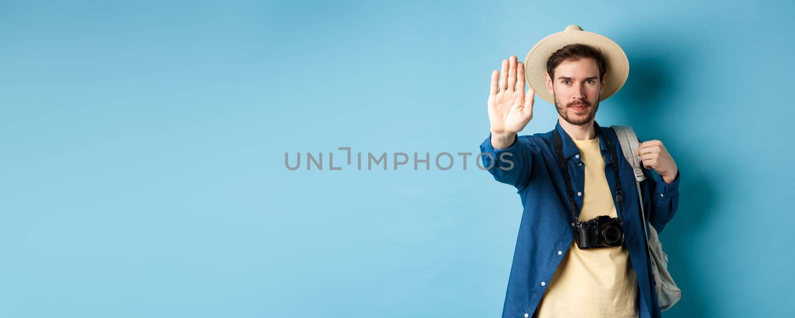 Serious male tourist warn you, stretch out hand to show stop gesture. Man on vacation forbid something, refusing or prohibiting action, standing with backpack on blue background.