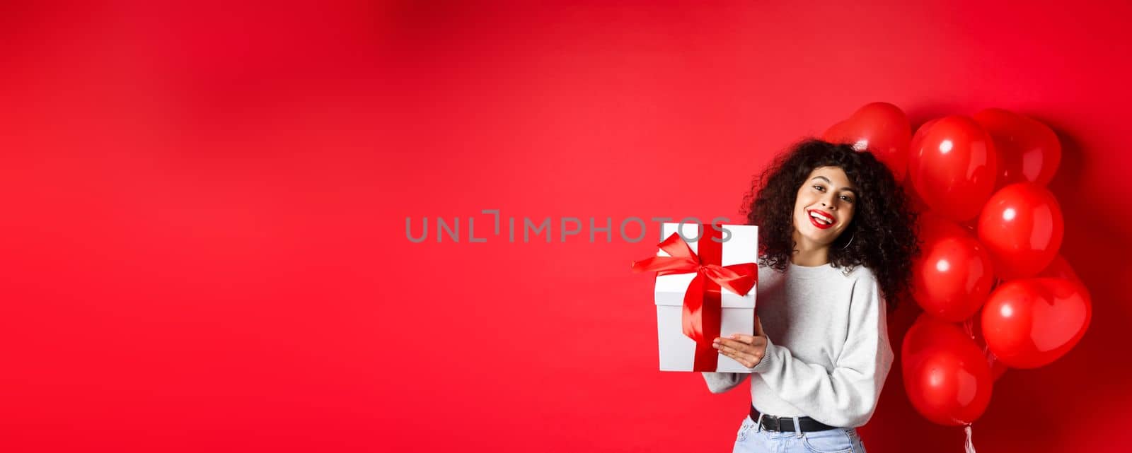 Holidays and celebration. Happy birthday girl holding gift and posing near party helium balloons, smiling excited at camera, red background by Benzoix
