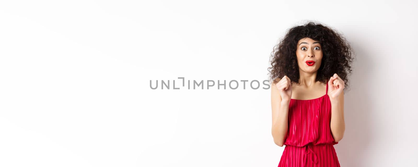 Excited smiling woman eager to try something, jumping from amusement and smiling, wearing red dress, standing on white background by Benzoix