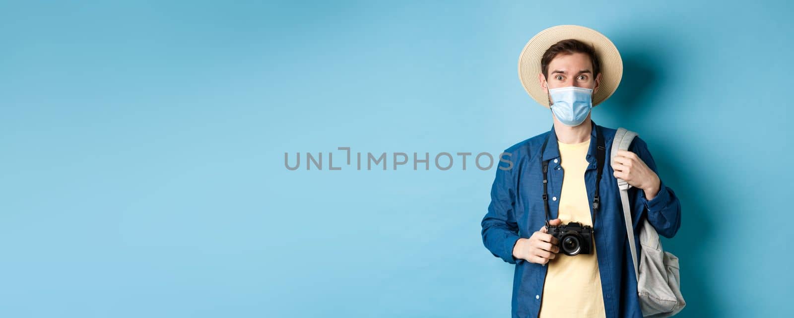 Covid-19 and travelling concept. Young guy tourist in medical mask and summer hat travel abroad during coronavirus pandemic, taking pictures on vacation, blue background by Benzoix