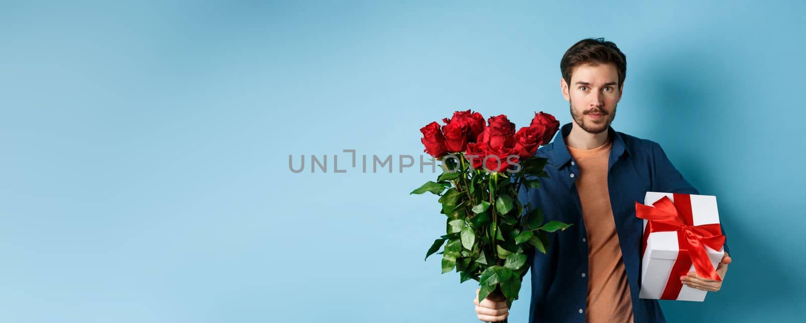 Romance and valentines day. Man presenting bouquet of red roses to lover. Boyfriend bring flowers and gift on romantic date, standing over blue background by Benzoix