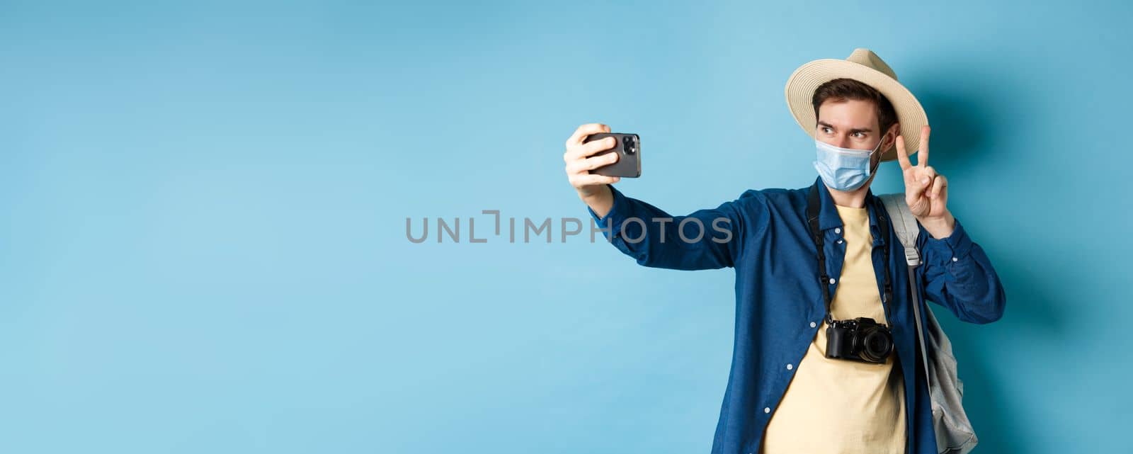 Covid-19, pandemic and travel concept. Happy and positive guy in tourist hat taking selfie and showing peace sign, posing near sightseeing, blue background by Benzoix