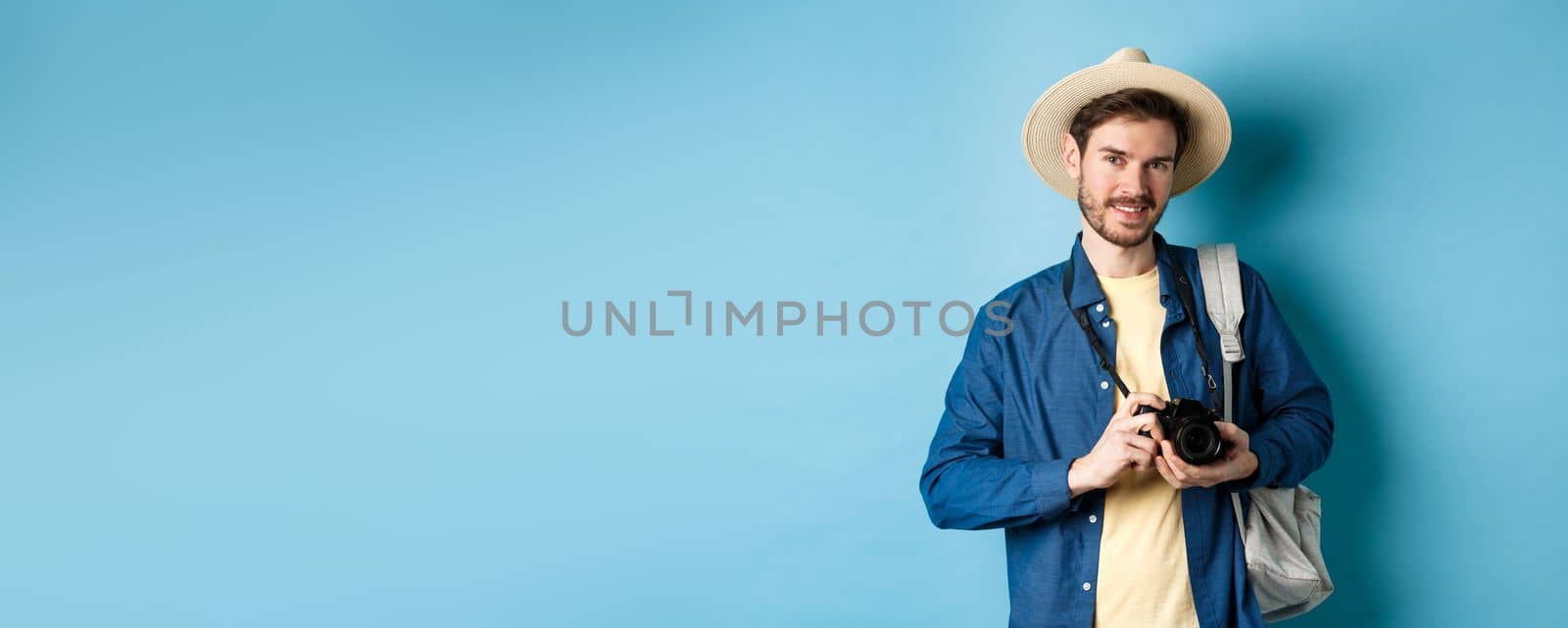 Handsome guy going on summer vacation, backpacking on holiday. Tourist with straw hat and camera smiling happy, standing on blue background.