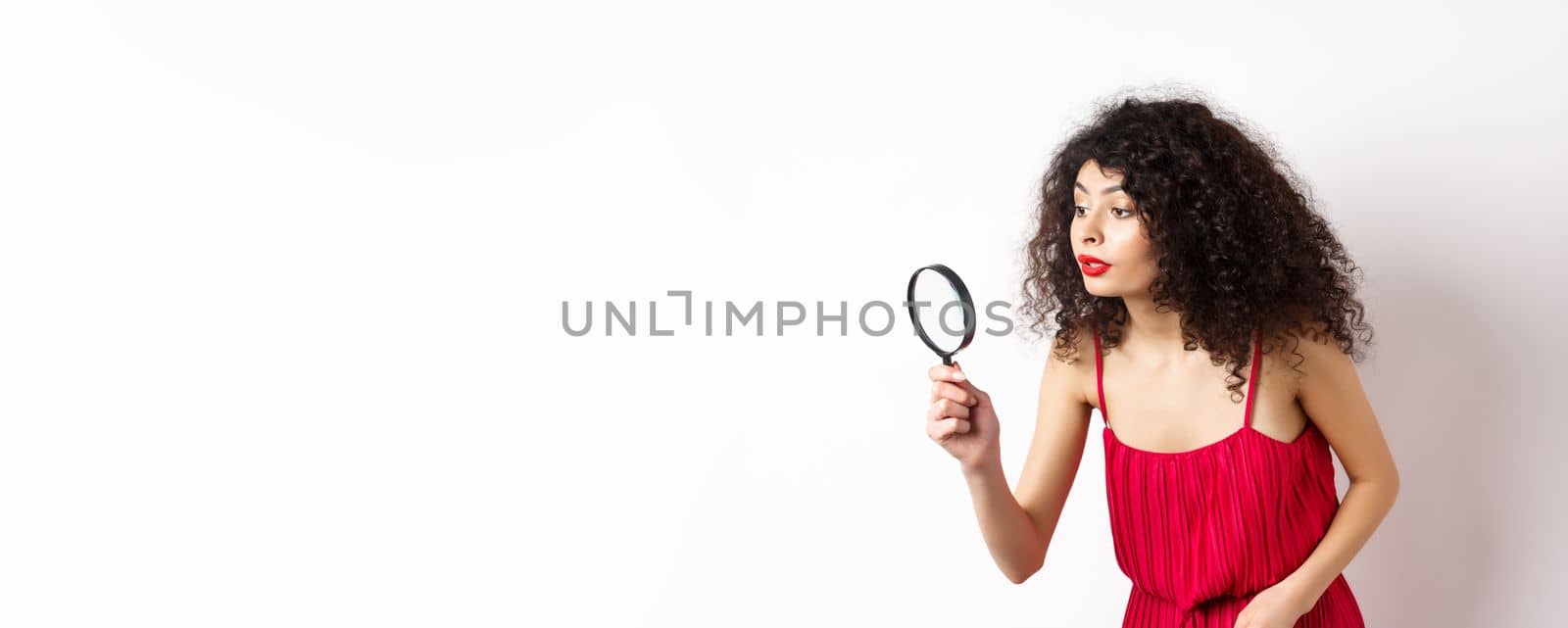 Beautiful woman in red dress searching for something, looking aside through magnifying glass, standing against white background.