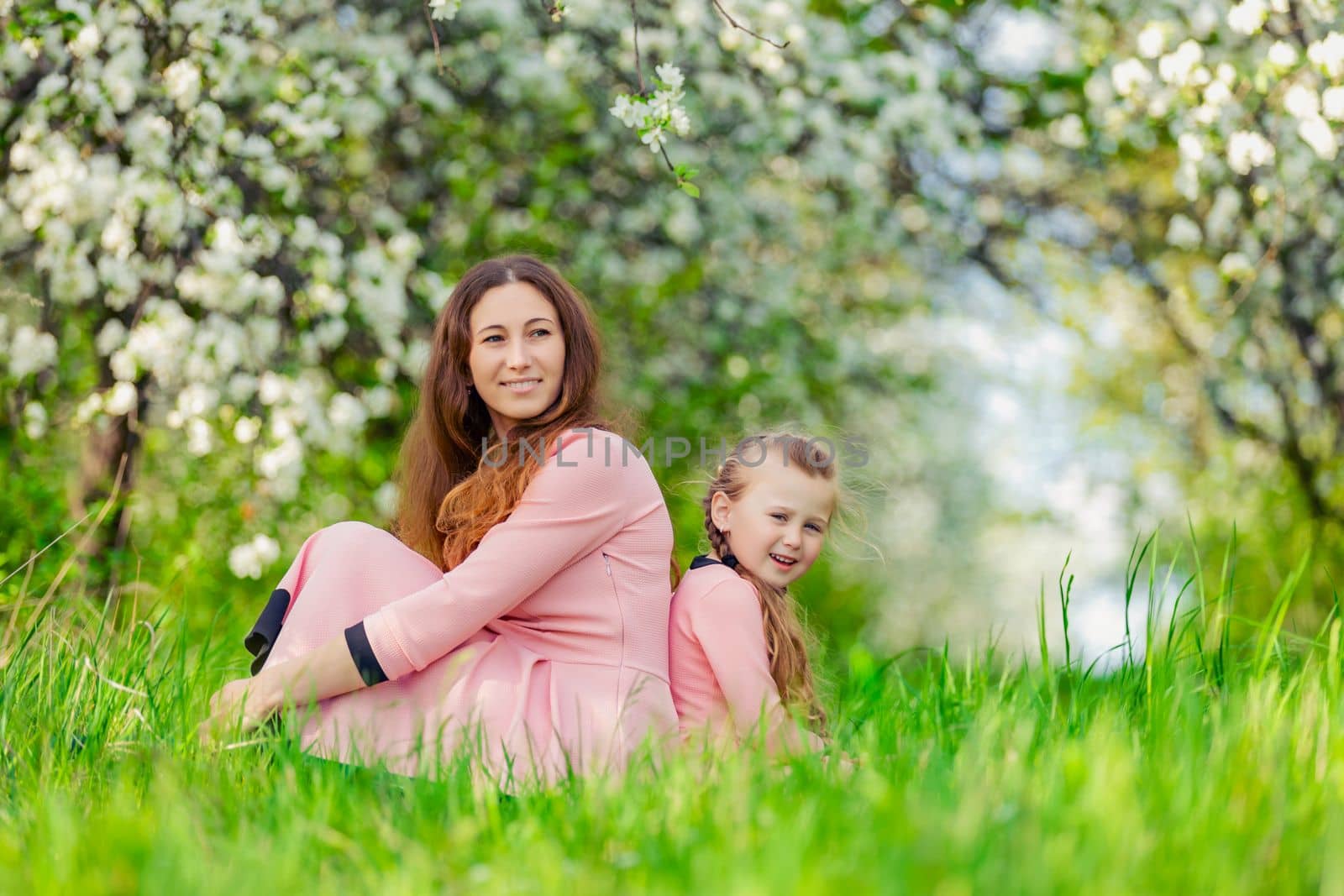 mother and daughter sit in a flowering garden with their backs to each other