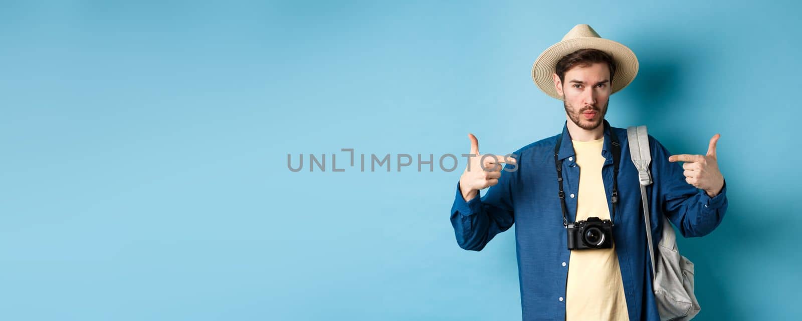 Confident and ready young man pointing at himself, self-promoting or show-off, going on summer vacation, wearing straw hat, holding backpack with camera, blue background.