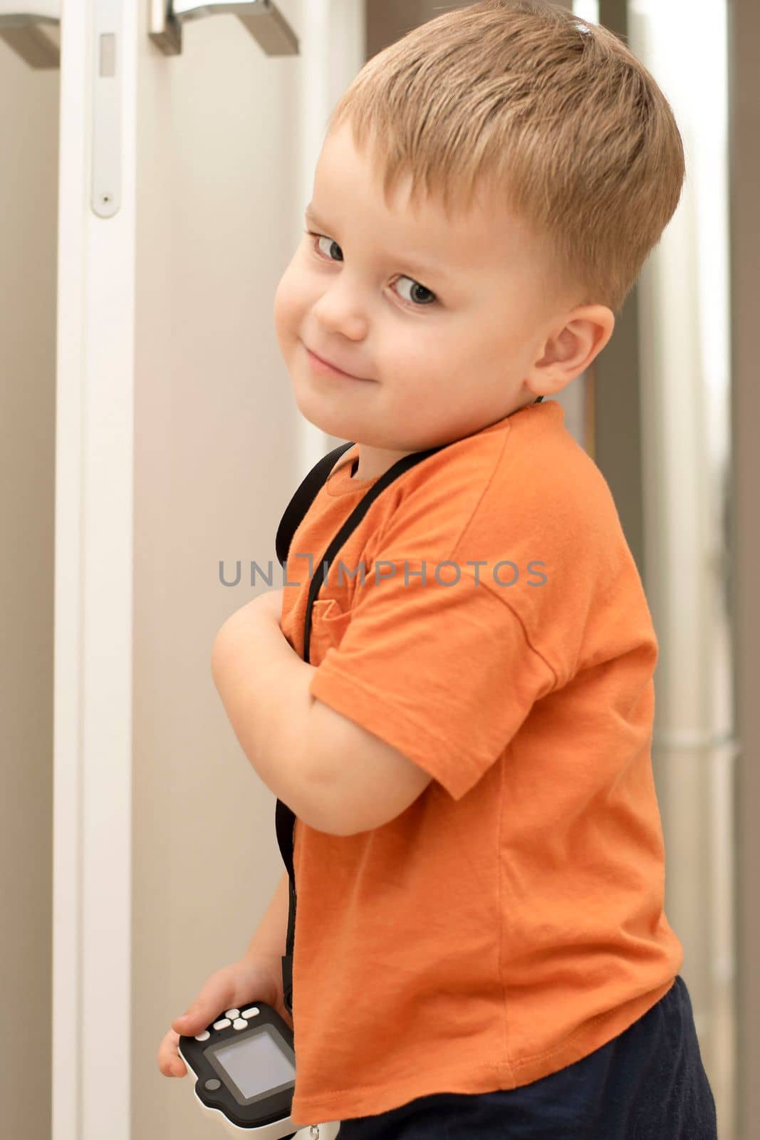 People. Handsome funny and emotional caucasian blond little boy in an orange t-shirt with a children's white camera takes pictures in the home interior. Close-up.