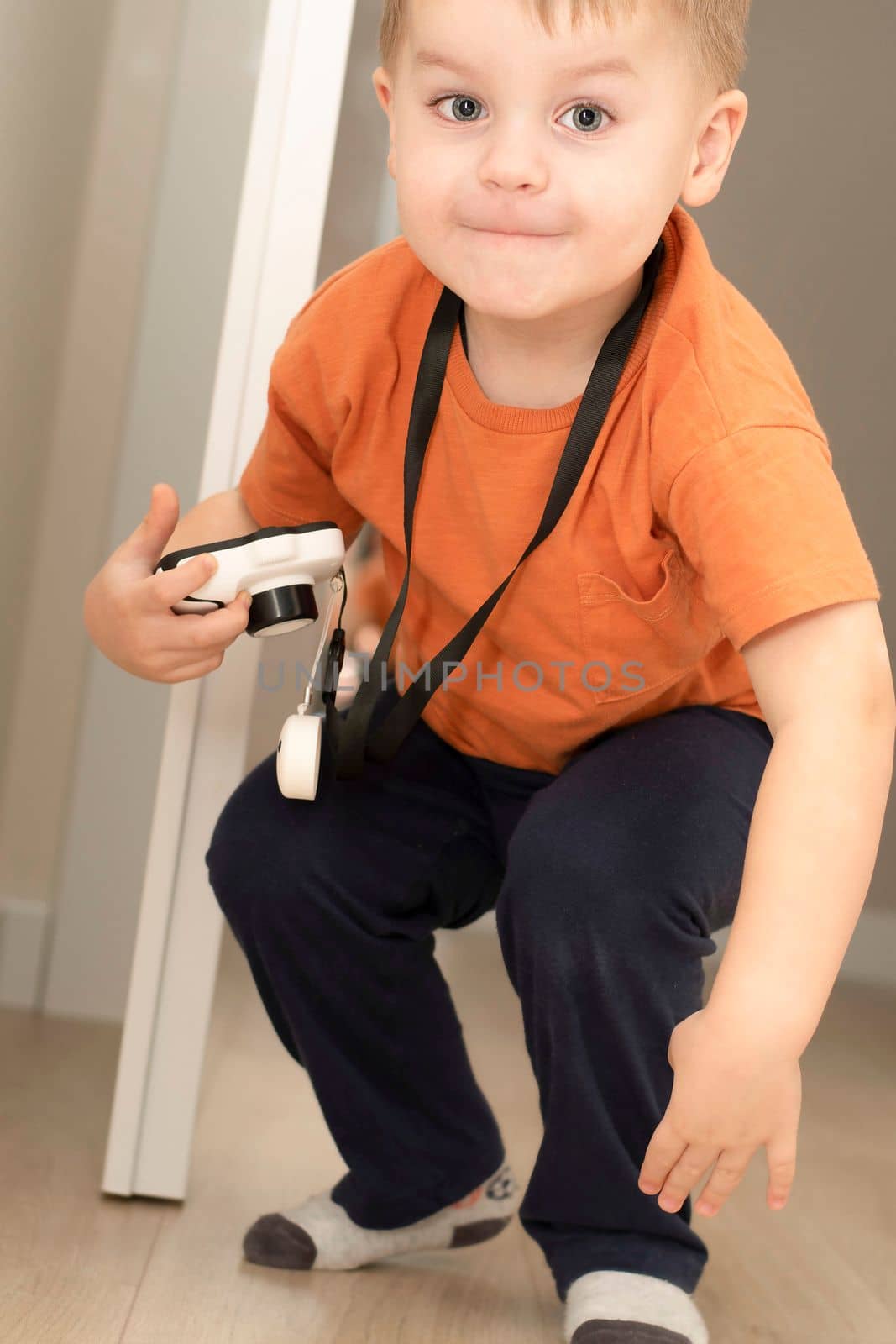 People. Handsome funny and emotional caucasian blond little boy in an orange t-shirt with a children's white camera takes pictures in the home interior. Close-up.