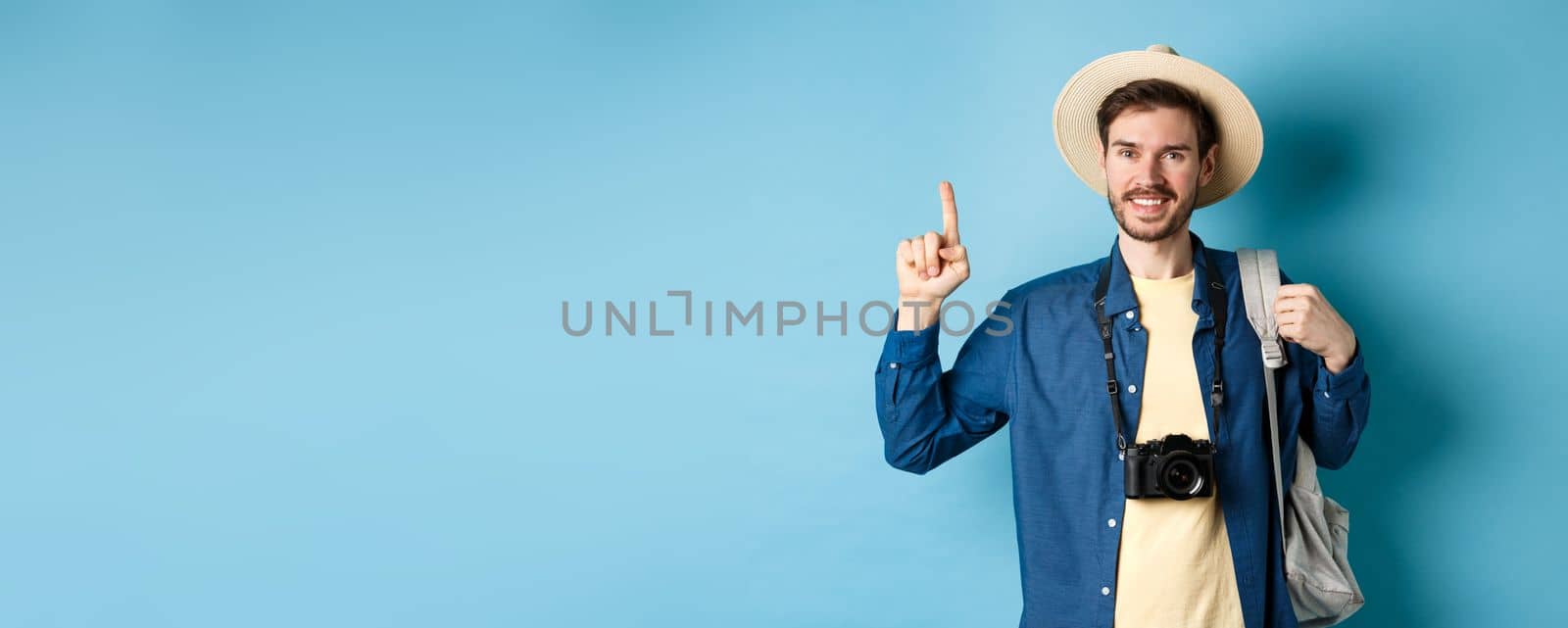 Handsome happy tourist in summer hat, holding backpack and camera, pointing finger up at logo, recommending travel agency or place on vacation, blue background by Benzoix