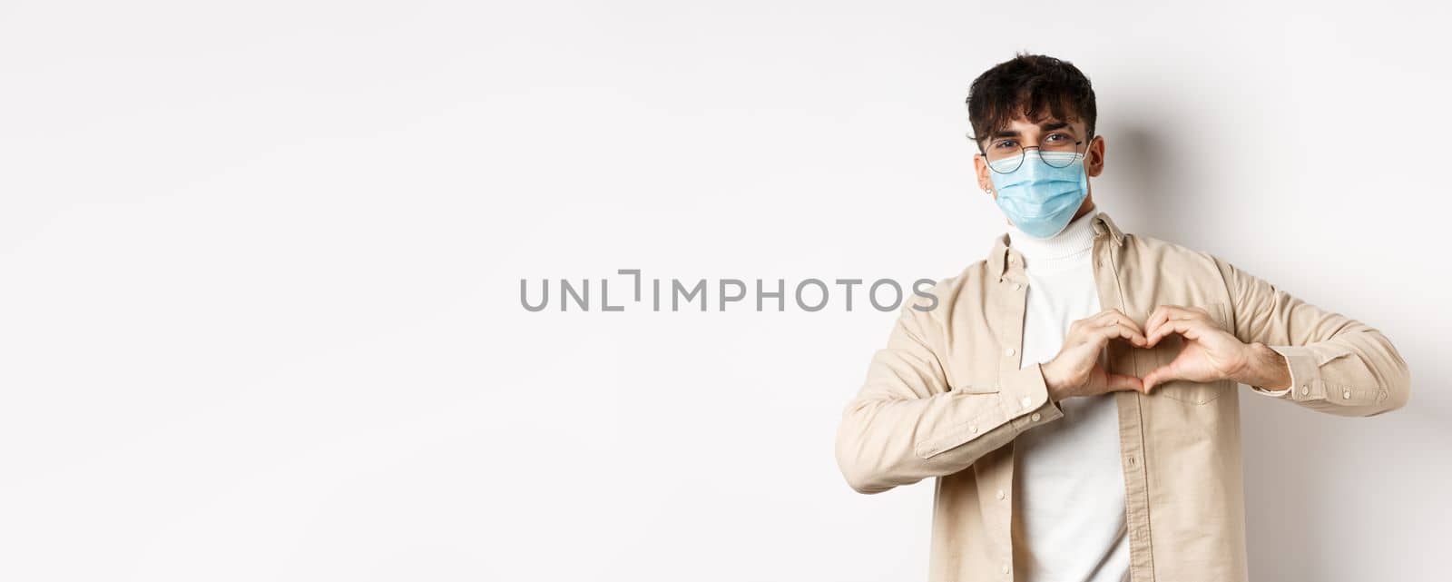 Health, covid and quarantine concept. Romantic young man in sterile medical mask showing heart gesture on chest, say I love you, standing on white background.