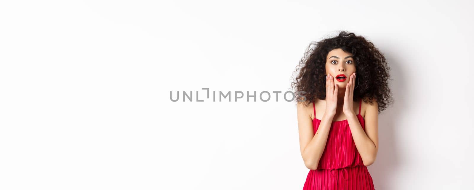 Excited curly-haired woman touch face with disbelief, looking amazed at camera, staring at promo, standing in red ress on white background by Benzoix