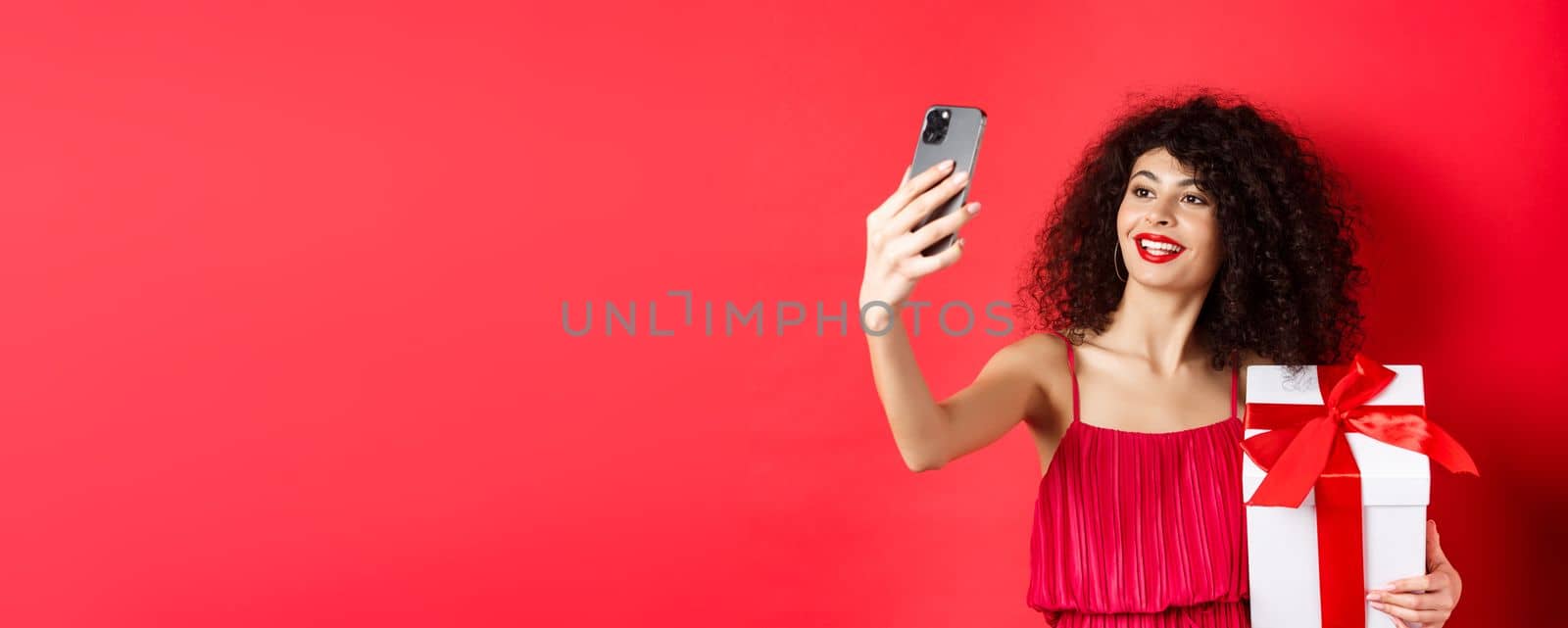 Beautiful girlfriend with curly hair, wearing evening dress, taking selfie with gift from lover, photographing on smartphone and smiling, standing over red background.