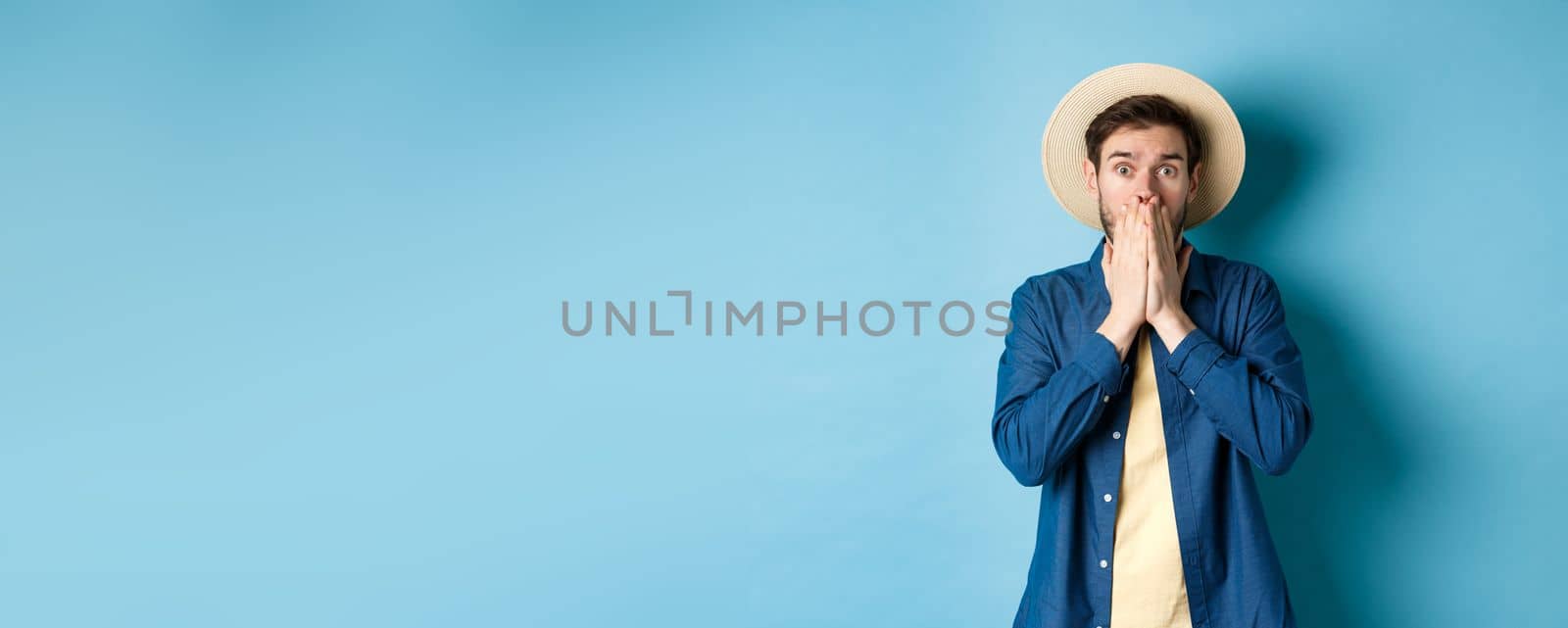 Shocked and alarmed tourist gasping, covering mouth with hands and looking startled at camera, standing on blue background by Benzoix